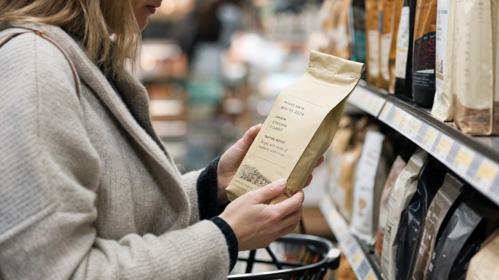 Shopper holding a bag of coffee in a supermarket aisle, reading the roast date and origin while shelves of coffee blur in the background.